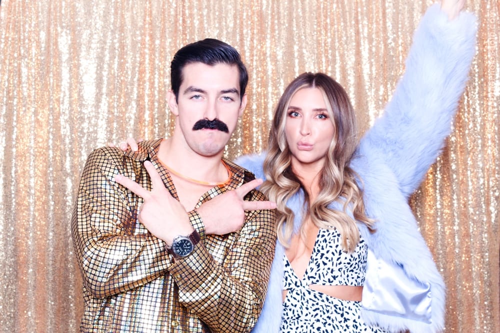 Couple posing in retro costumes with a gold sequined backdrop, showcasing 70s-themed party vibes and playful expressions.