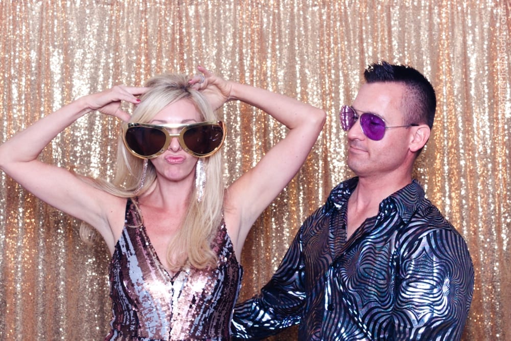 Couple posing playfully in oversized sunglasses against a shimmering gold backdrop, embodying a fun, retro vibe suitable for a Studio 54-themed party.