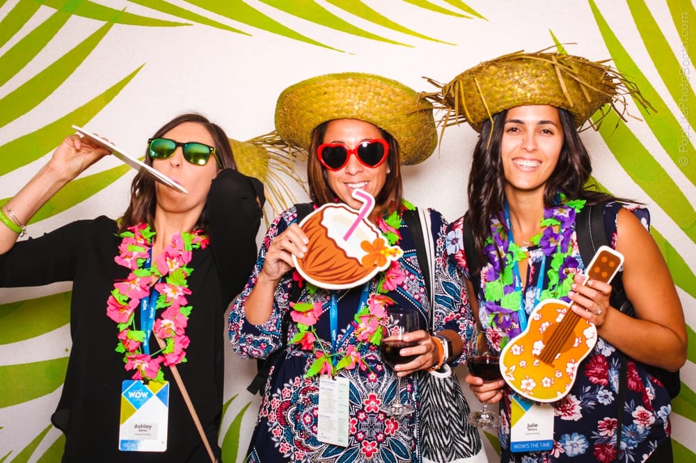 Three women in tropical attire with leis and straw hats posing in a photo booth, holding fun props like a coconut drink and a ukulele, against a vibrant palm leaf backdrop, capturing the festive spirit of a luau-themed party.