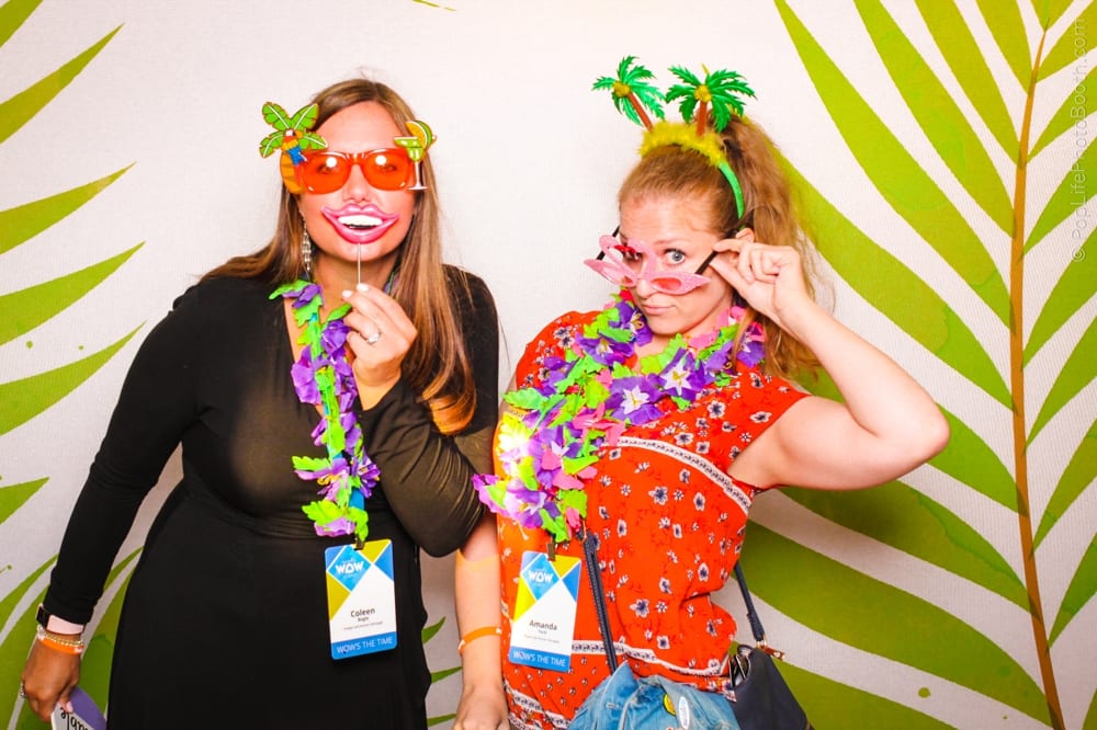 Two women wearing tropical-themed attire and props, including leis and oversized sunglasses, posing playfully in front of a vibrant palm leaf backdrop, capturing the festive spirit of a luau-themed party.