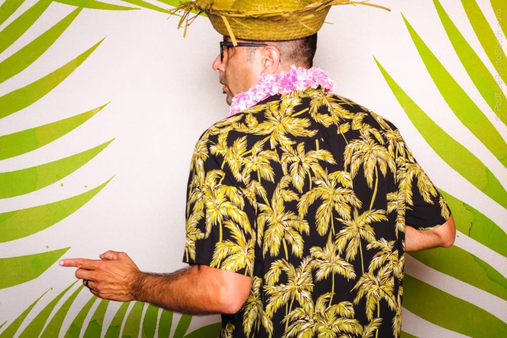 Man wearing a tropical shirt and straw hat with a floral lei, posing in front of a green palm leaf backdrop, evoking a festive luau atmosphere.