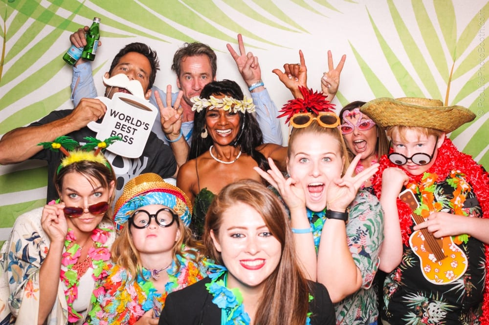Group of people in tropical attire with leis and props, posing joyfully in a photo booth, featuring a backdrop of palm leaves, capturing a festive luau party theme.