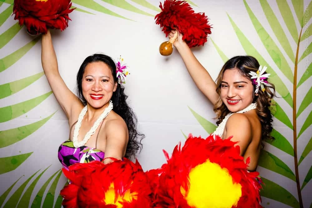 Two women in vibrant Hawaiian attire holding large red pom-poms and a gourd, posing joyfully against a tropical backdrop, embodying the festive spirit of a Tropical Luau-themed party.