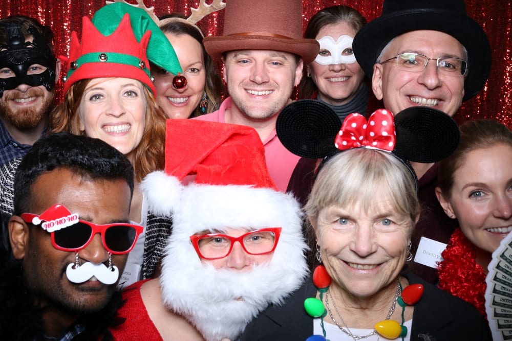 Group of people in festive attire, including Santa hats, elf ears, and holiday-themed props, posing together in front of a shiny red backdrop, capturing the fun spirit of a company holiday party with a photo booth.