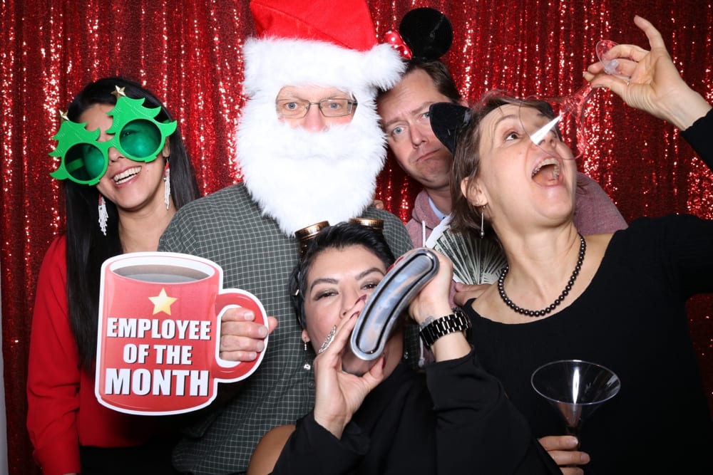 Group of friends celebrating in a photo booth with festive props, including a Santa hat, green holiday glasses, and an "Employee of the Month" sign, against a shiny red backdrop.