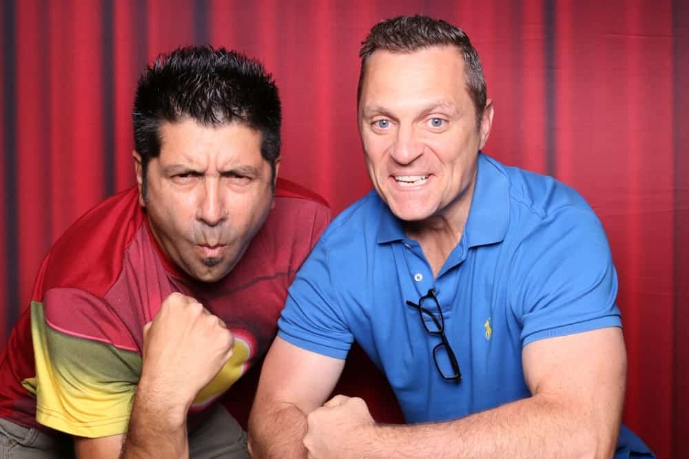 Two men posing playfully in a photo booth with a red backdrop, showcasing fun expressions and strong poses, ideal for capturing memorable moments at a corporate party or event.