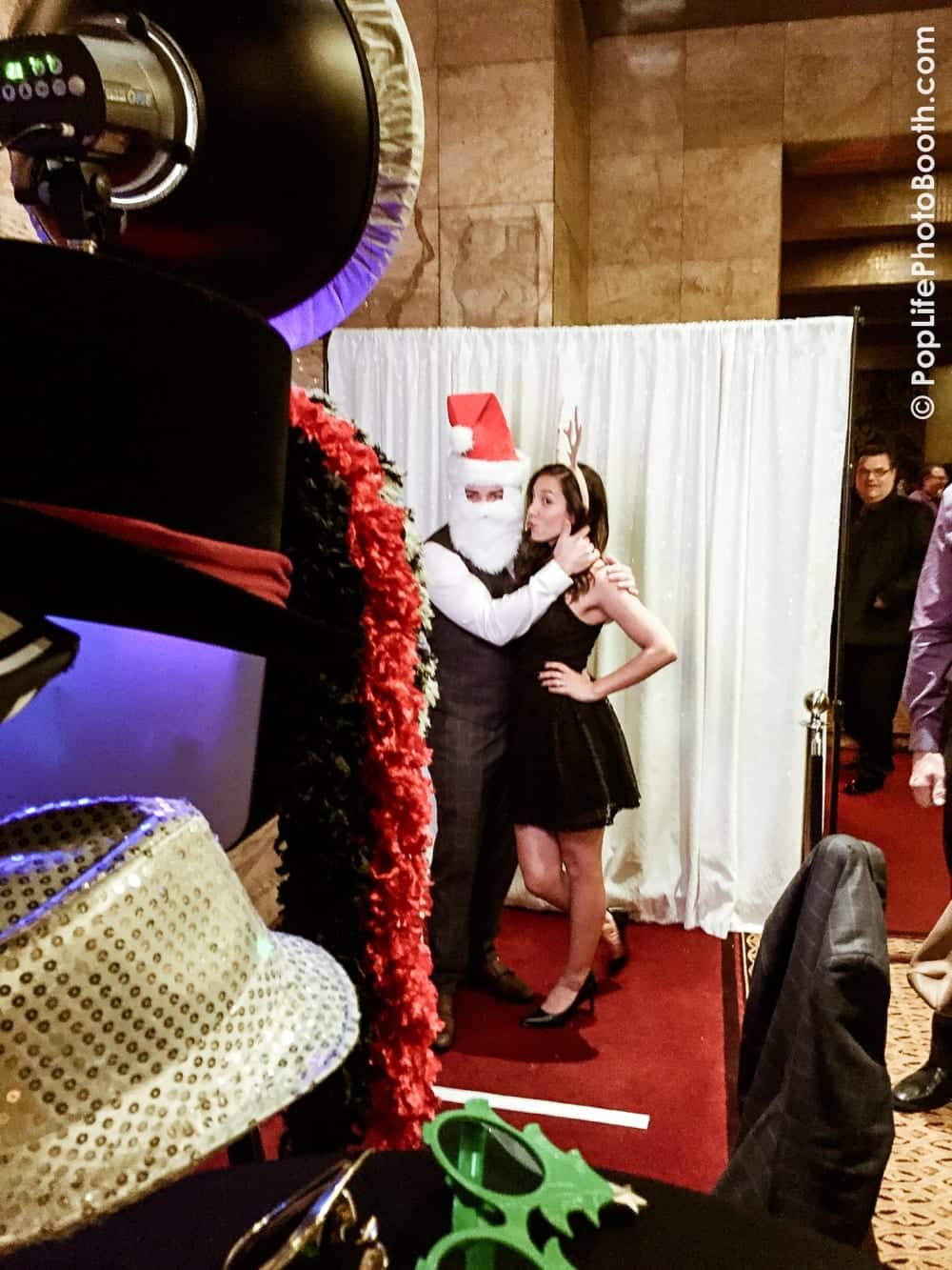 Santa Claus posing with a woman in a festive setting, featuring a photo booth backdrop, holiday decorations, and props like a Santa hat and reindeer antlers, capturing the spirit of a corporate holiday party.