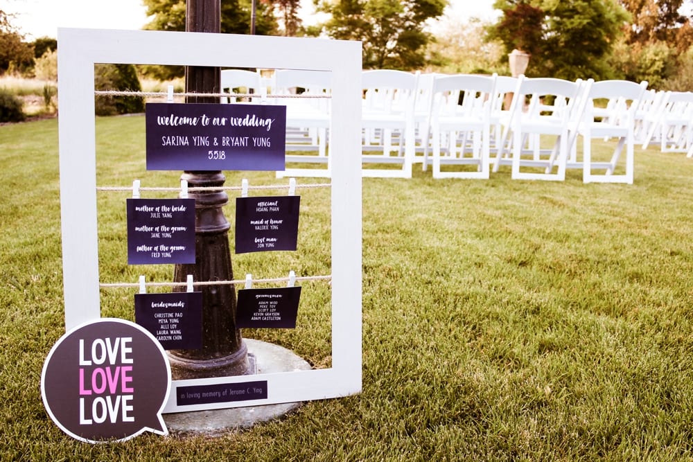 Wedding welcome sign displaying names and roles of family members, surrounded by white chairs on a grassy outdoor venue, with a decorative "Love" sign in memory of a loved one.