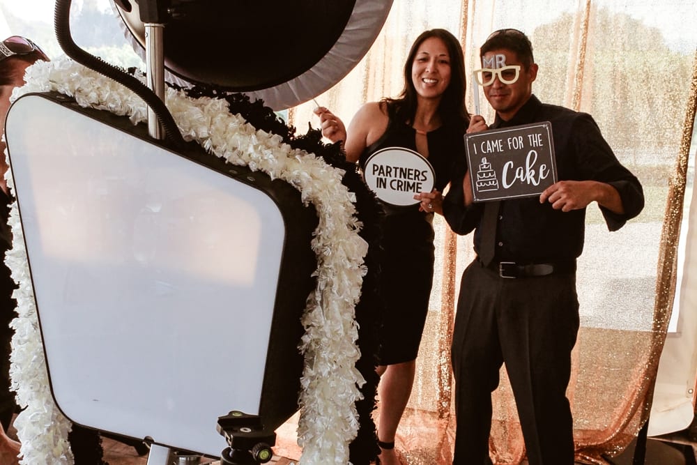 Couple posing with playful signs at a wedding photo booth, featuring "Partners in Crime" and "I Came for the Cake," surrounded by floral decorations, emphasizing fun and celebration.