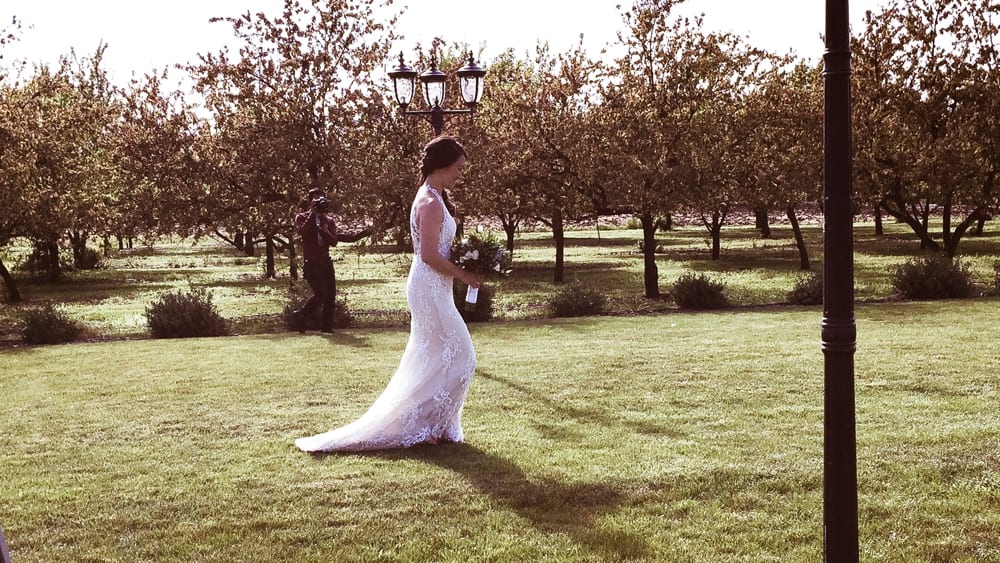 Bride in elegant lace wedding dress walking in a garden setting, surrounded by trees and greenery, with a photographer capturing the moment.