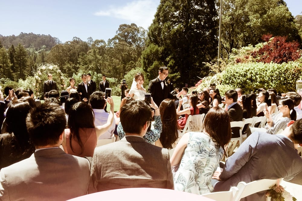 Wedding ceremony scene with bride and groom standing at altar, surrounded by wedding party and guests, capturing moments on smartphones, lush greenery in background.