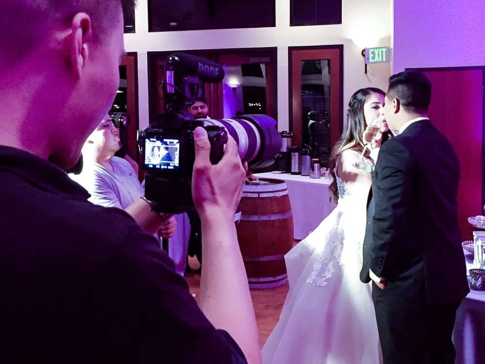 Wedding couple sharing a moment during reception, photographer capturing the scene, festive atmosphere with purple lighting, wedding decor in background.