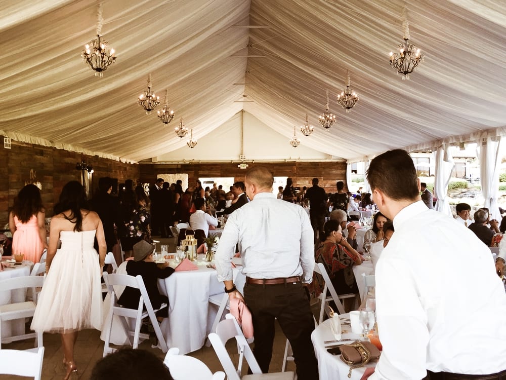 Wedding reception scene with guests mingling and dining under a tent, featuring tables set with elegant decor and chandeliers, capturing the celebratory atmosphere of a wedding event.