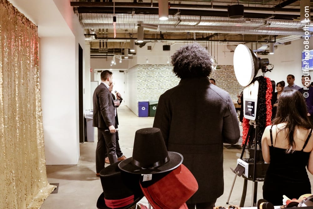 Guests enjoying a corporate Thanksgiving event with a photo booth setup, featuring a gold sequin backdrop, props including hats, and attendees engaging in conversation.