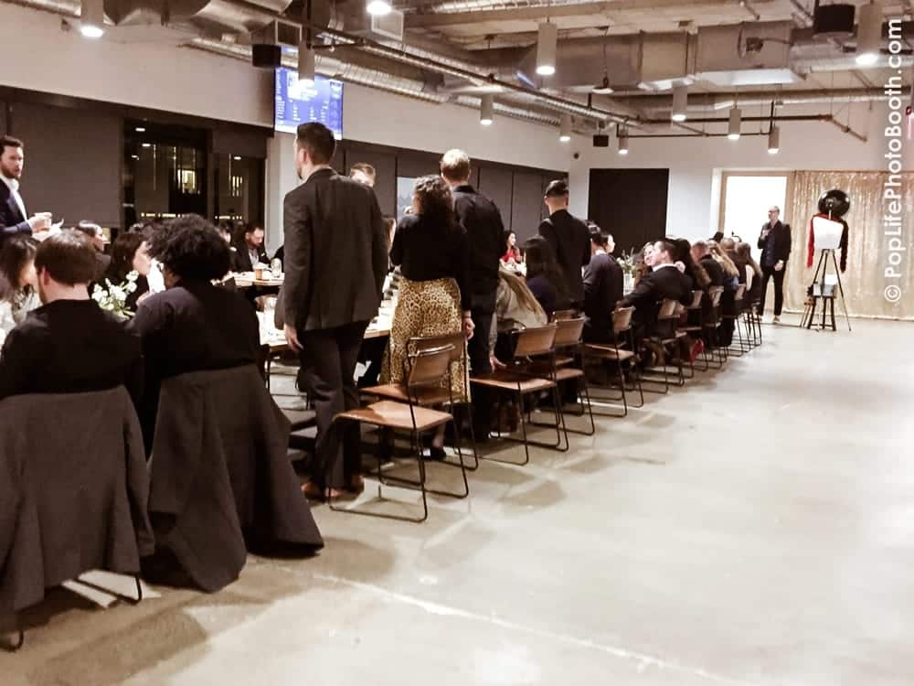Guests enjoying a Thanksgiving corporate event at a long dining table, with a photo booth setup in the background, highlighting the festive atmosphere provided by Pop Life Photo.