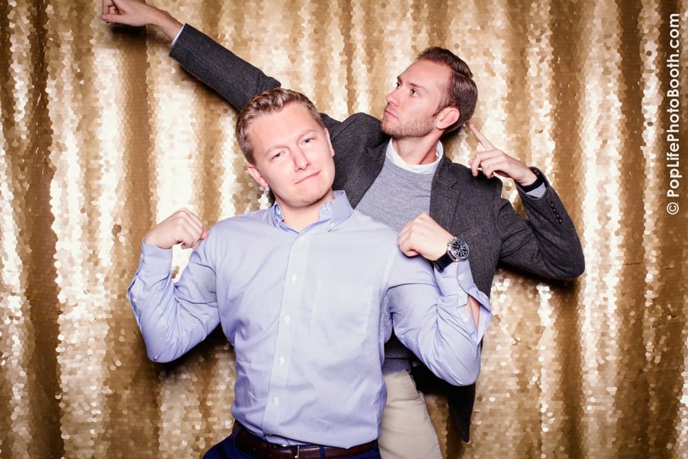 Two men posing playfully in a photo booth with a shimmering gold backdrop, one flexing muscles and the other pointing upwards, capturing the fun atmosphere of a corporate Thanksgiving event hosted by Yelp with Pop Life Photo.