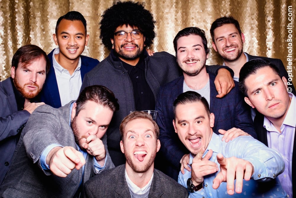 Group of men posing playfully in a photo booth at a Thanksgiving corporate event, featuring festive attire and a glittery backdrop, showcasing camaraderie and fun during the celebration.