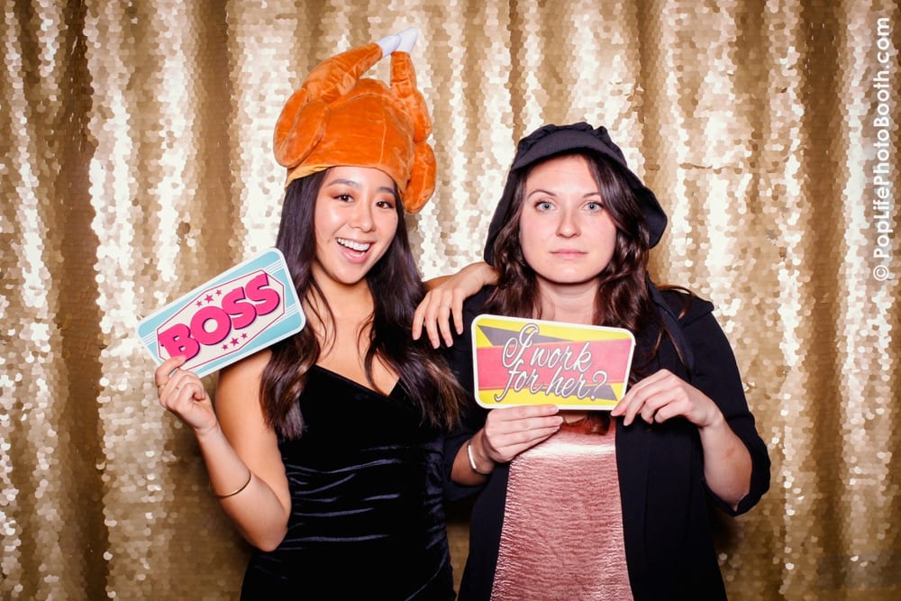 Two women posing in a photo booth with Thanksgiving-themed props, one wearing a turkey hat and holding a sign that says "BOSS," while the other holds a sign reading "I work for her?" with a sparkling golden backdrop.