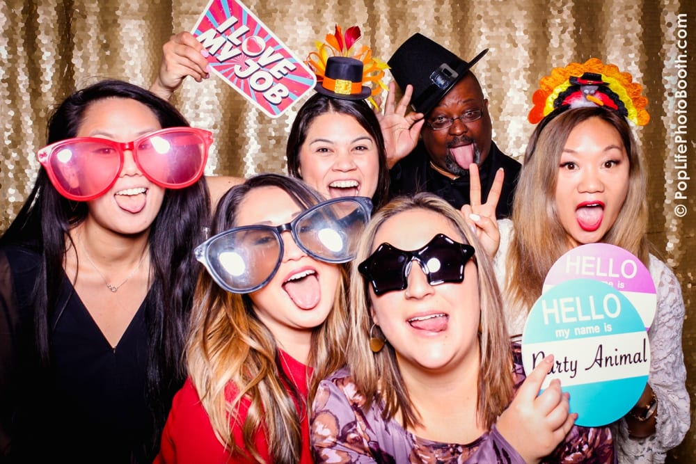 Group of guests at a Thanksgiving corporate event wearing fun props, including oversized glasses and hats, posing playfully in a photo booth with a sparkling backdrop.