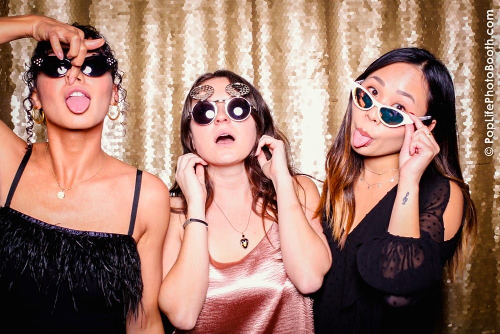 Three women posing playfully in sunglasses at a photo booth, surrounded by a shimmering gold backdrop, capturing the festive spirit of a Thanksgiving corporate event hosted by Yelp.