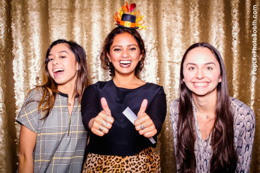 Three women posing joyfully in a photo booth, wearing fun Thanksgiving props, including a turkey hat, against a shimmering gold backdrop, capturing the festive spirit of a corporate Thanksgiving event hosted by Yelp.