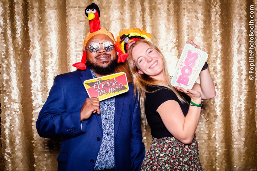 Guests at a Thanksgiving-themed photo booth wearing festive props, including a turkey hat and sunglasses, holding signs that read "I work for her?" and "BOSS," with a sparkling gold backdrop.