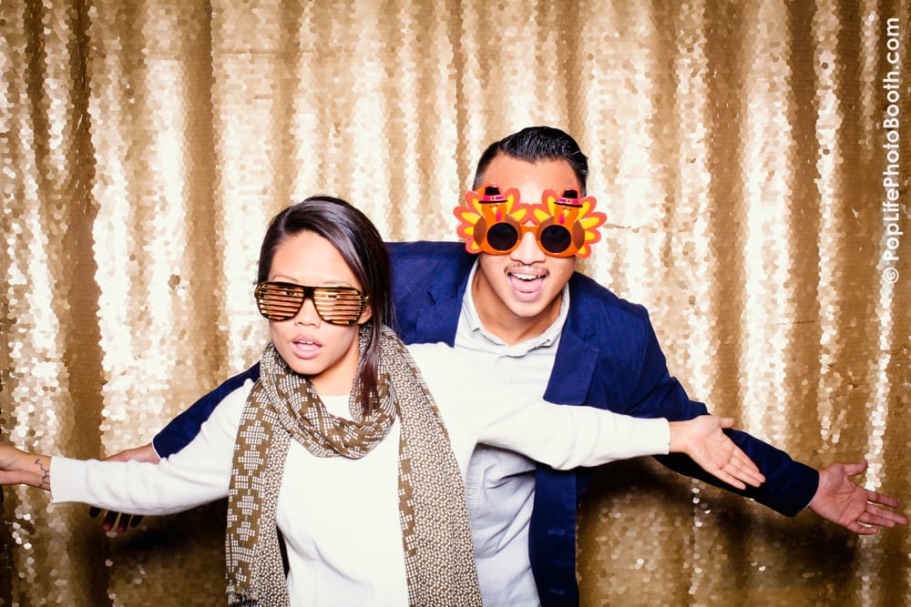 Guests enjoying a photo booth at a Thanksgiving event, wearing festive props like turkey-themed glasses and oversized sunglasses, with a shimmering gold backdrop.
