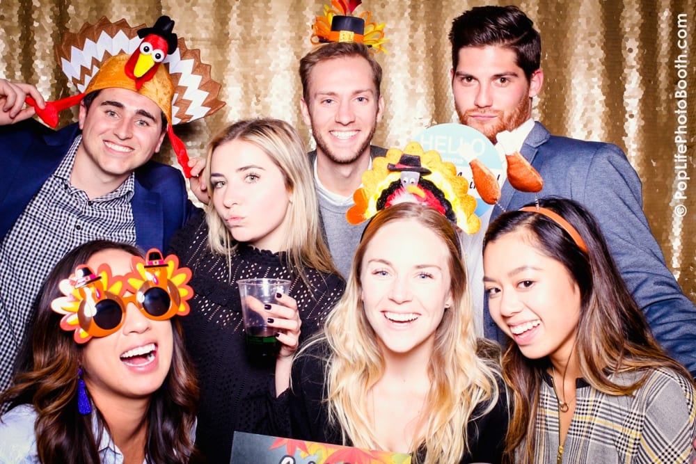 Group of young adults wearing Thanksgiving-themed props, including turkey hats and festive glasses, enjoying a photo booth at a corporate event hosted by Yelp, with a sparkling gold backdrop.