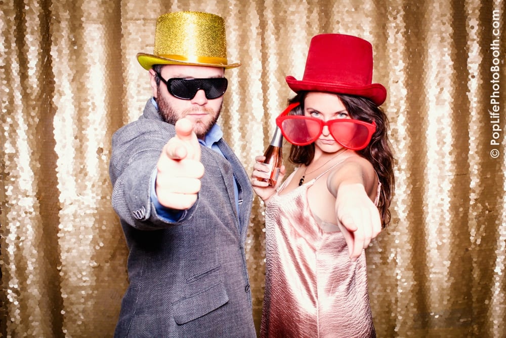 Guests at a Thanksgiving corporate event posing in a photo booth, wearing a gold hat, sunglasses, and a red hat with oversized glasses, against a sparkling backdrop.