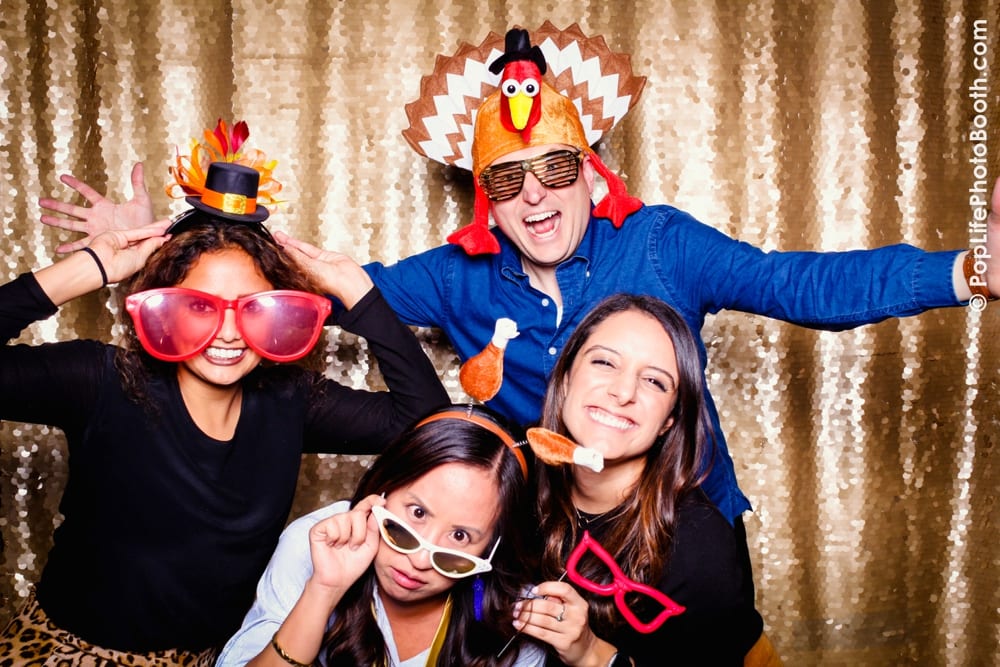 Four guests enjoying a Thanksgiving-themed photo booth, wearing fun props like oversized glasses, a turkey hat, and a small top hat, with a sparkling gold backdrop.