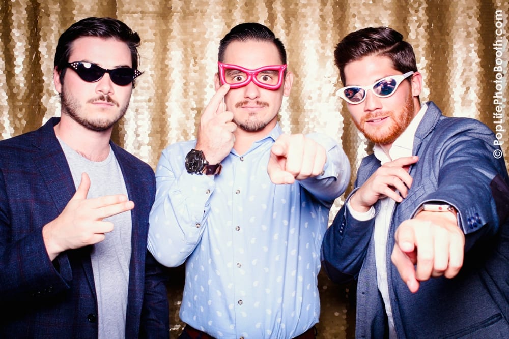 Three men posing playfully in a photo booth with fun props, including oversized sunglasses, against a shimmering gold backdrop, capturing the festive spirit of a Thanksgiving corporate event hosted by Yelp.