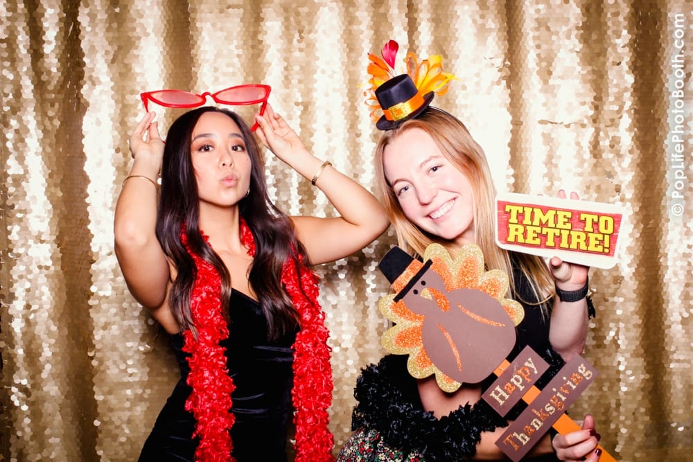 Two women posing playfully in a photo booth, wearing festive Thanksgiving props including oversized glasses, a turkey hat, and signs reading "Happy Thanksgiving" and "Time to Retire," with a sparkling gold backdrop.