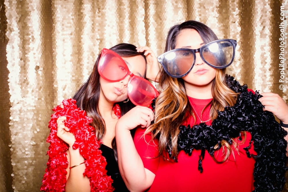 Two women posing playfully in a photo booth, wearing oversized novelty glasses and colorful feather boas, with a shimmering gold backdrop, capturing the festive spirit of a Thanksgiving corporate event hosted by Yelp.