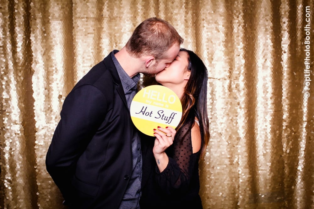Couple kissing in a photo booth, holding a sign that reads "HELLO, my name is Hot Stuff," with a sparkling gold backdrop, capturing festive moments from a Thanksgiving corporate event.