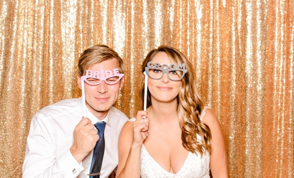 Bride and groom wearing playful glasses with "BRIDE" and "GROOM" text, posing in front of a shimmering gold backdrop at a wedding photo booth.