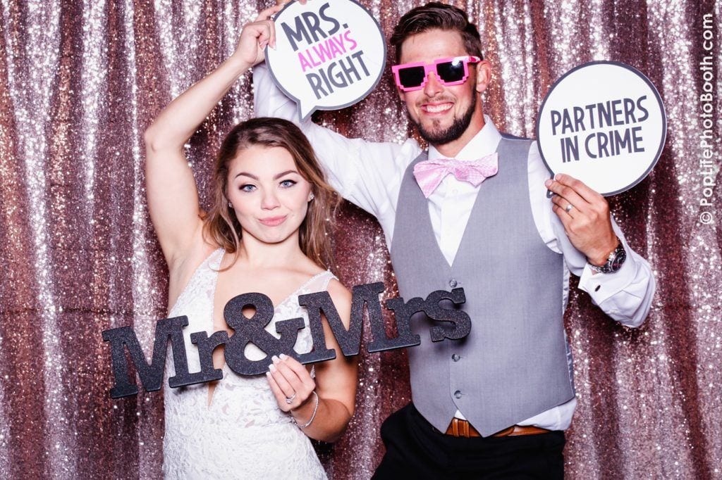 Couple posing in wedding attire with "Mr & Mrs" sign and playful props, including "Mrs. Always Right" and "Partners in Crime," against a sparkling backdrop, celebrating their wedding at Pop Life Photo Booth.