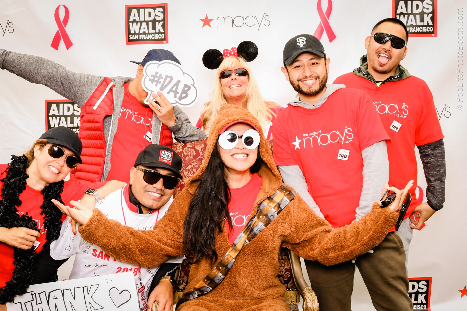 Group of diverse participants at the SFAids Walk in San Francisco, wearing red event shirts and playful costumes, posing cheerfully with props in front of a branded backdrop featuring the event logo and sponsor Macy's.