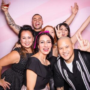 Group of six friends posing joyfully in a vintage-inspired photo booth, showcasing retro fashion and playful expressions, ideal for themed parties and events.