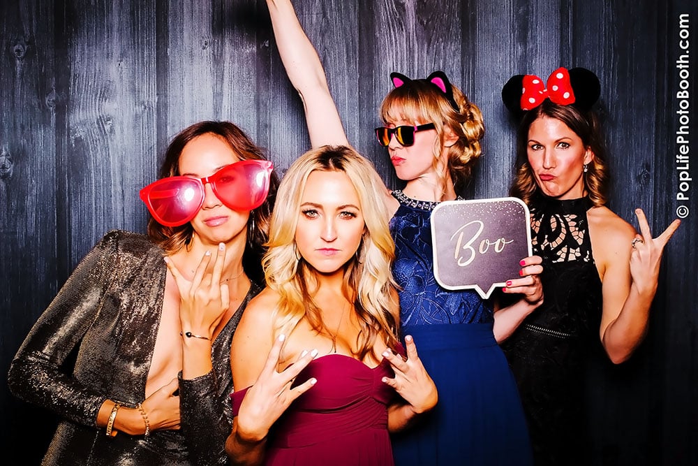Four women posing in a photo booth at a wedding, wearing fun accessories like oversized sunglasses and playful headbands, with one holding a sign that says "Boo," against a dark wooden backdrop.