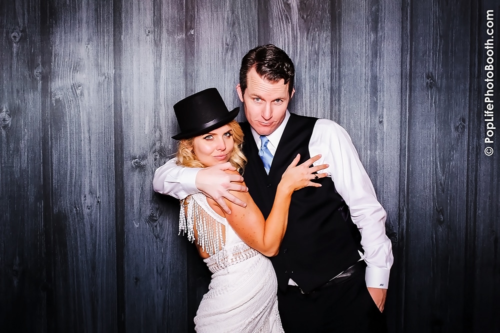 Happy newly married couple posing playfully in a photo booth, with the bride wearing a sparkling gown and a black hat, capturing joyful wedding memories at Terra Gallery, San Francisco.