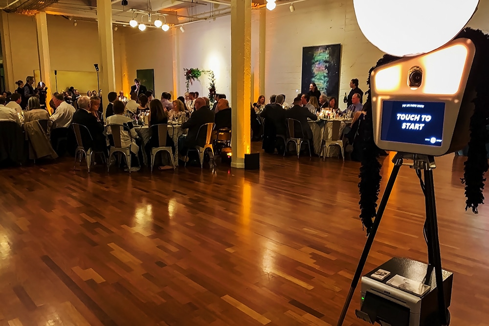 Interior view of Terra Gallery in San Francisco, featuring guests at elegantly set tables and a photo booth with a "Touch to Start" screen, highlighting its suitability for weddings and corporate events.