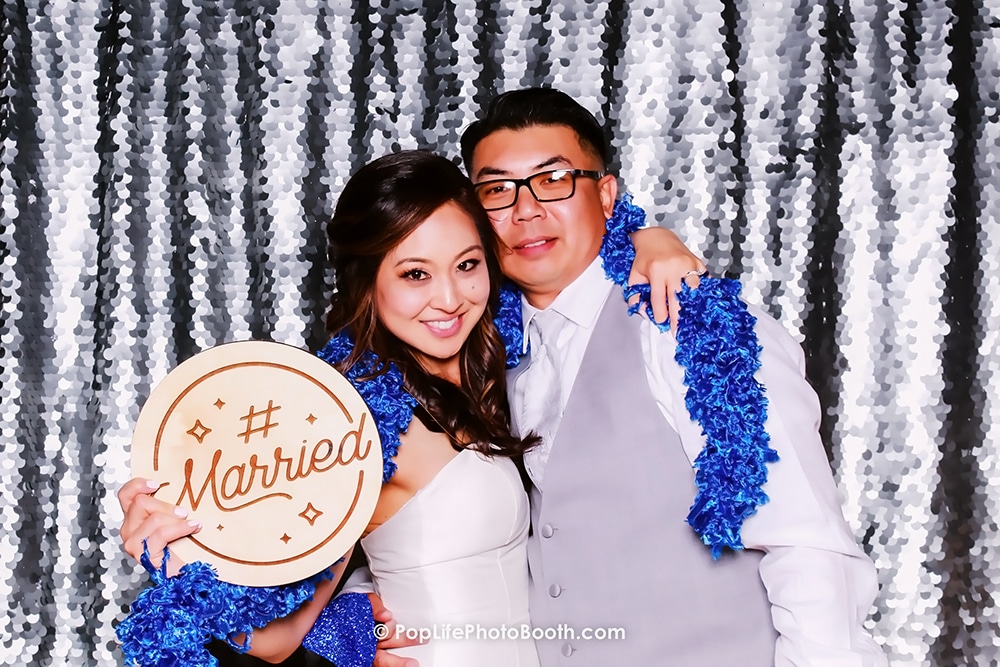 Just married couple holding a "#Married" sign, smiling in front of a silver sequins backdrop at Casa Real, Livermore, celebrating their wedding.