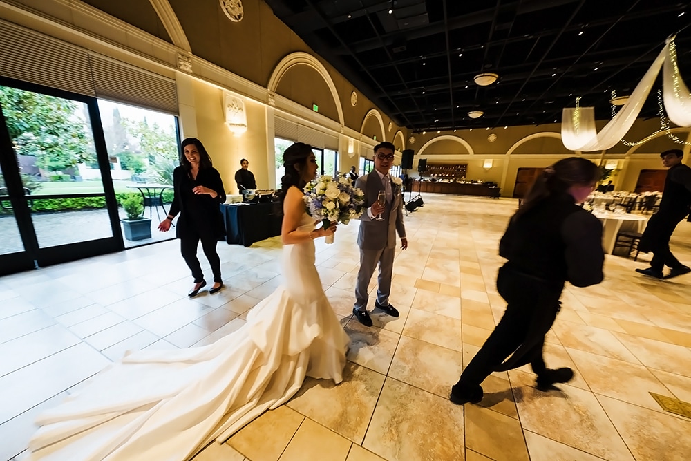 Bride in elegant wedding dress holding bouquet, smiling, with guests and staff in Casa Real venue, showcasing spacious event space and decor.