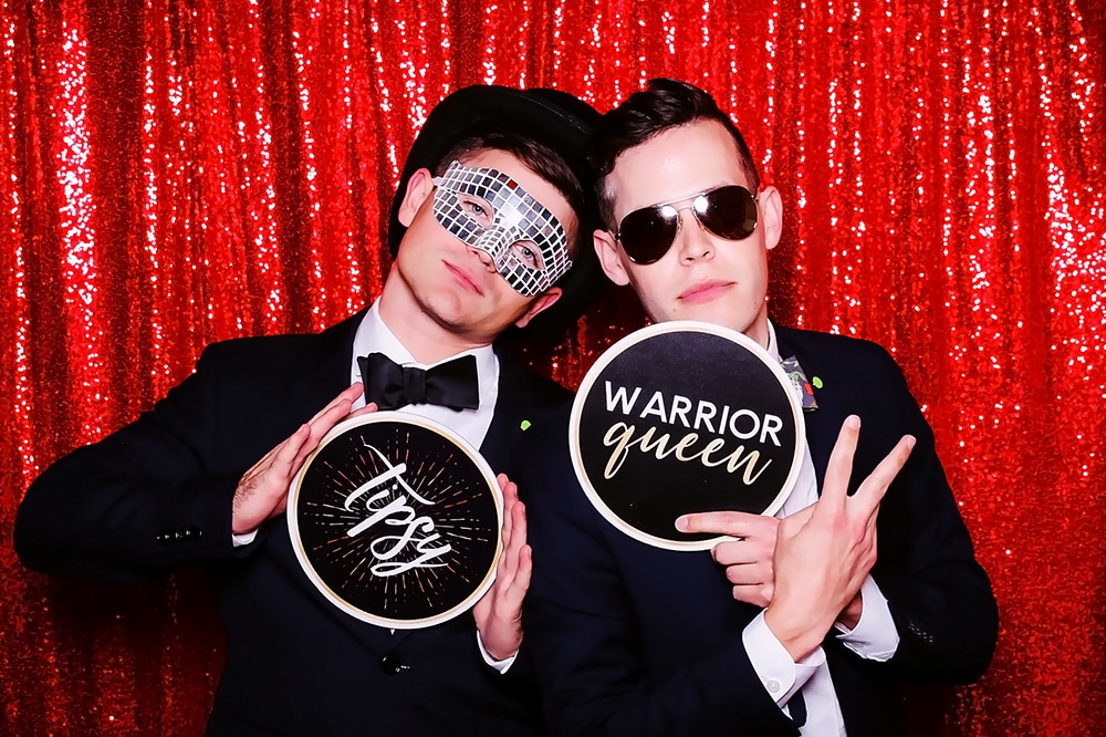 Two men in formal attire posing in a photo booth, one wearing a mirrored disco mask and holding a sign that says "Tipsy," while the other wears sunglasses and holds a sign that reads "Warrior Queen," against a sparkling red backdrop, capturing the fun and festive spirit of celebrations.