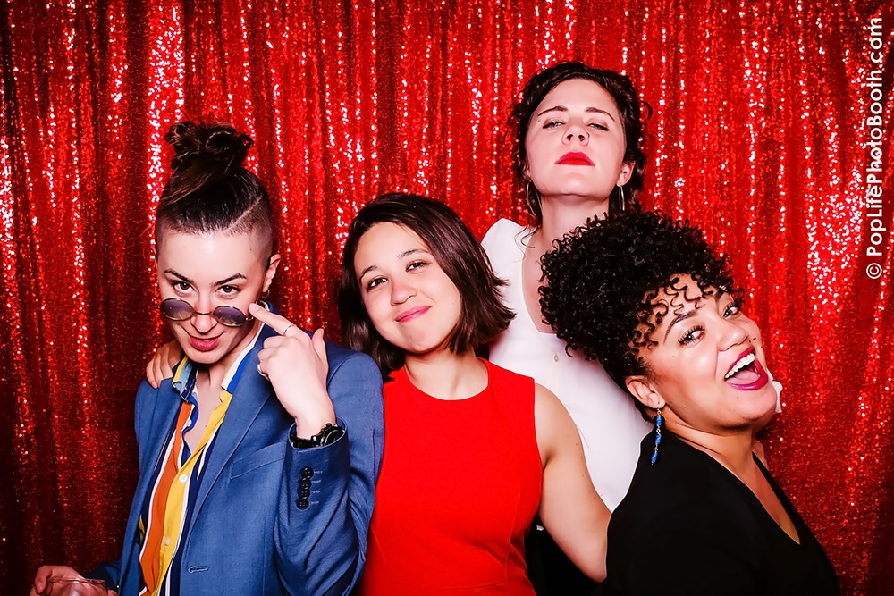 Group of four friends posing playfully in a photo booth with a shiny red backdrop, showcasing joy and celebration, ideal for event memories.