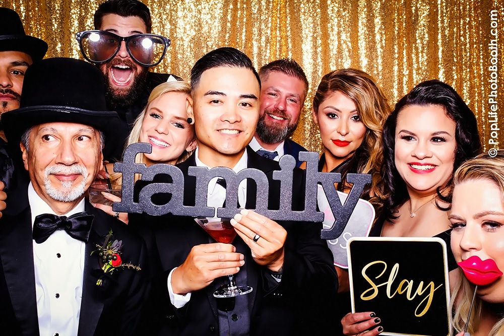 Group photo booth shot at a wedding, featuring diverse guests holding signs that read "family" and "Slay," with a sparkling gold backdrop, showcasing the fun atmosphere of The General Residence venue in San Francisco.