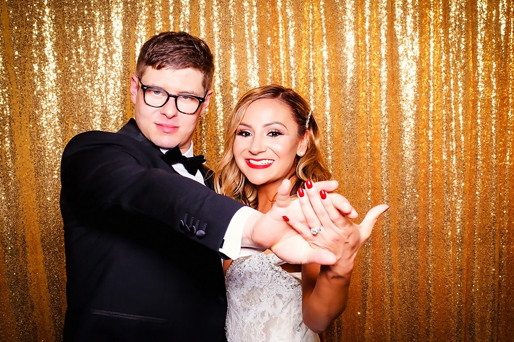 Newlywed couple posing in a photo booth with a sparkling gold backdrop, showcasing wedding attire and joyful expressions, emphasizing the celebratory atmosphere of events in San Francisco.