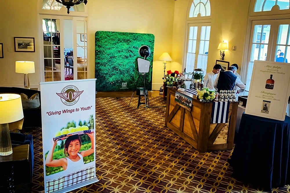 Photo booth setup at The Olympic Club in San Francisco, featuring a green backdrop, a smiling child on a promotional banner, and a bar area with staff preparing drinks.