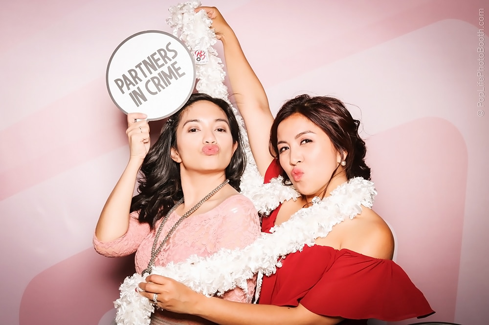 Two women posing playfully in a photo booth, one holding a sign that says "PARTNERS IN CRIME," adorned with leis, in front of a pink backdrop, capturing a fun moment at an event.