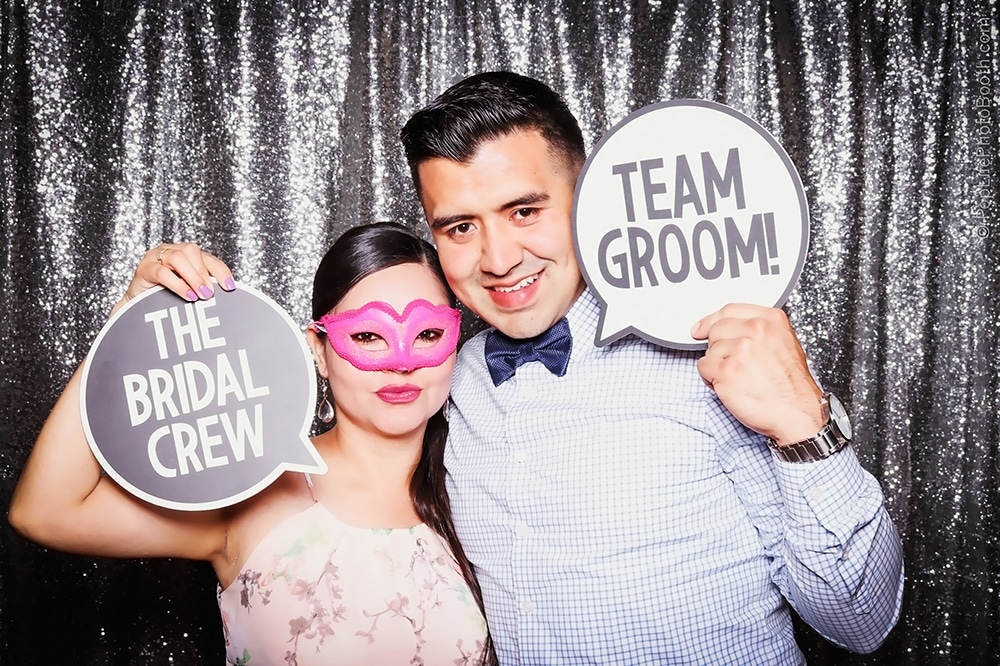 Couple posing in a photo booth at a wedding, holding signs that read "THE BRIDAL CREW" and "TEAM GROOM!" in front of a shimmering backdrop, capturing a fun moment at an event in Livermore.